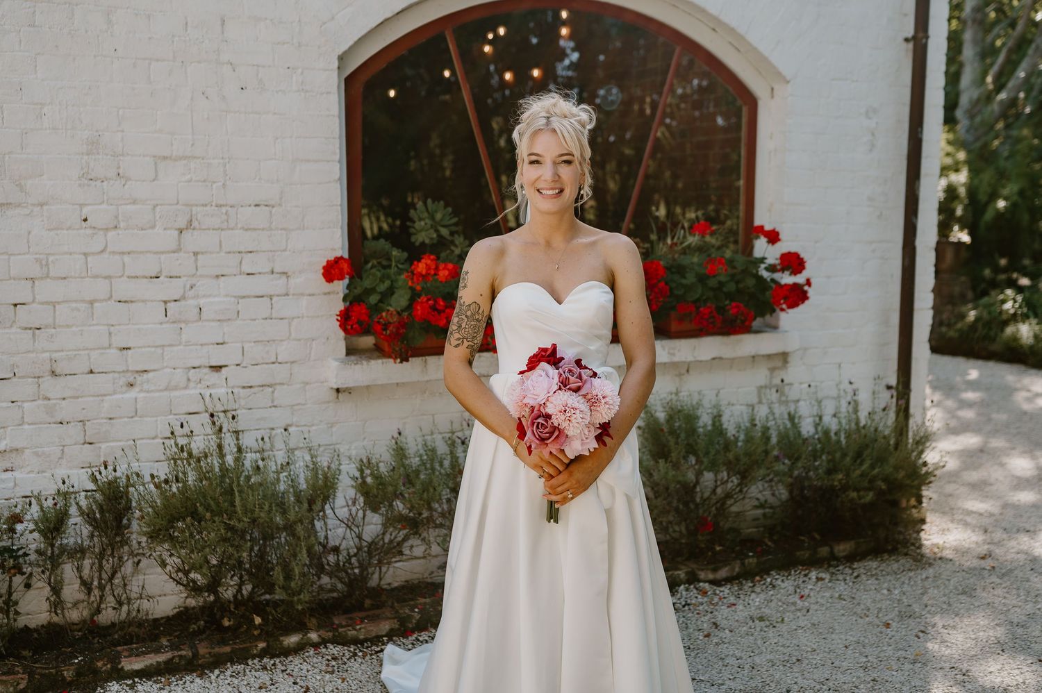 A bride wearing a classic white strapless wedding gown holds a pink bouquet in front of a white wall with red flowers.