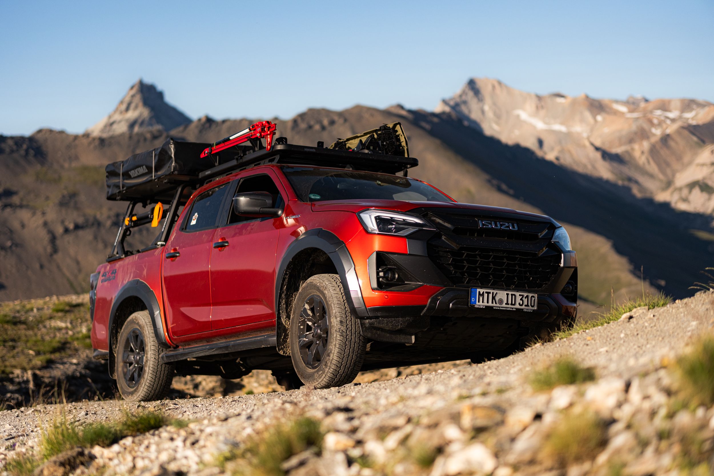 Red off-road pickup truck traversing rugged mountain terrain against a backdrop of rocky peaks and blue sky.