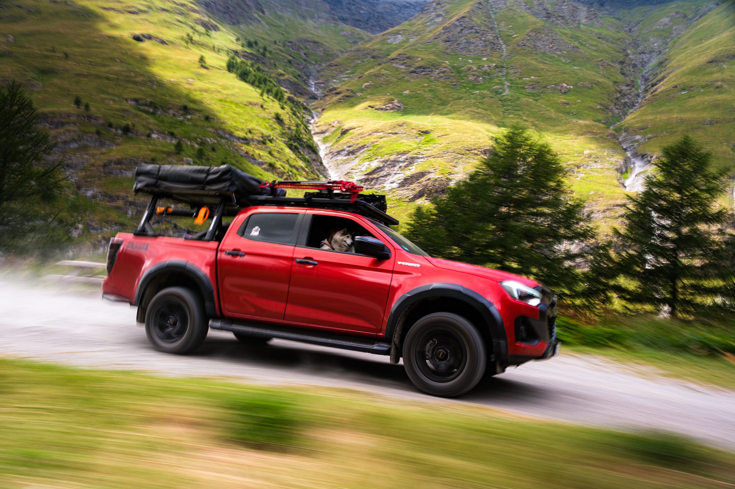 A red pickup truck drives along a scenic mountain road with lush green hills and valleys in the background.