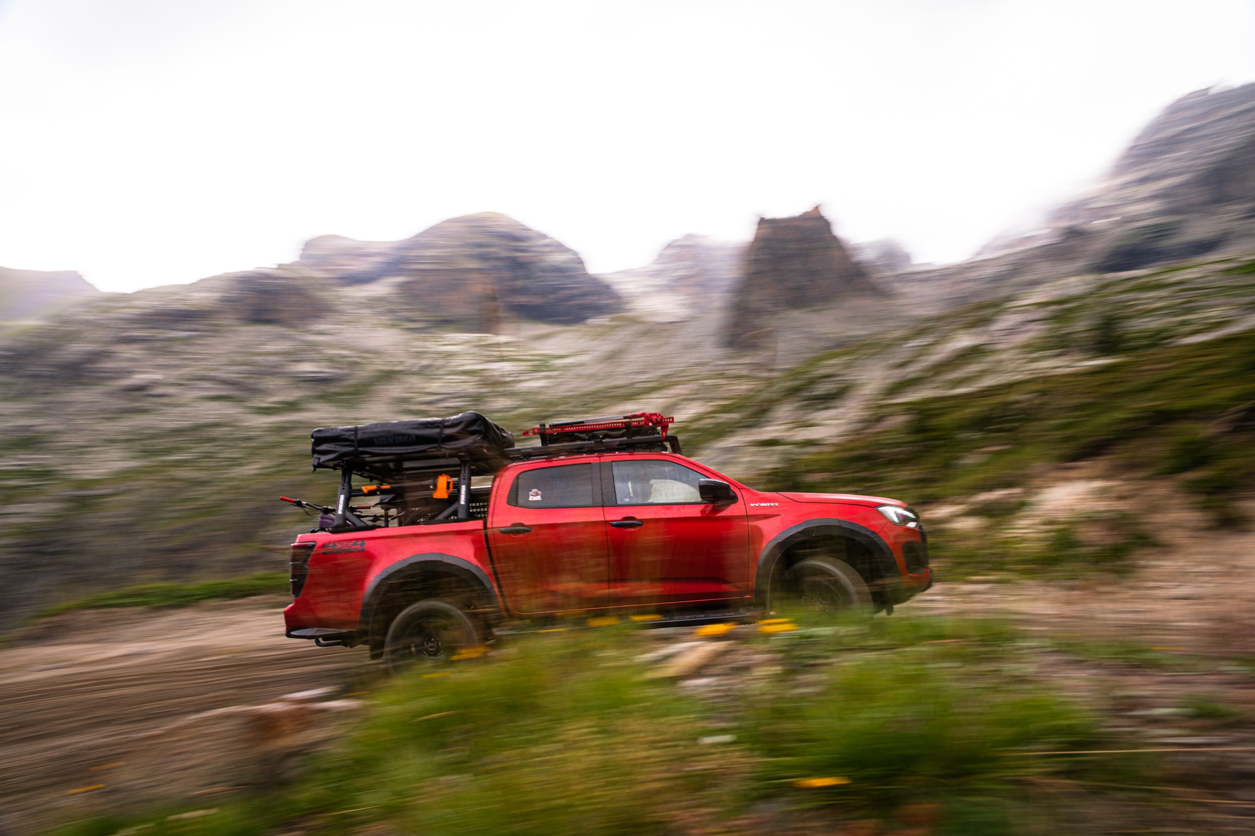 A red pickup truck drives along a winding mountain road through rugged desert terrain and steep cliffs.