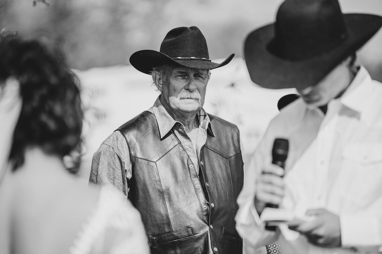 Black and white candid image showing figures in western attire with cowboy hats conversing outdoors.