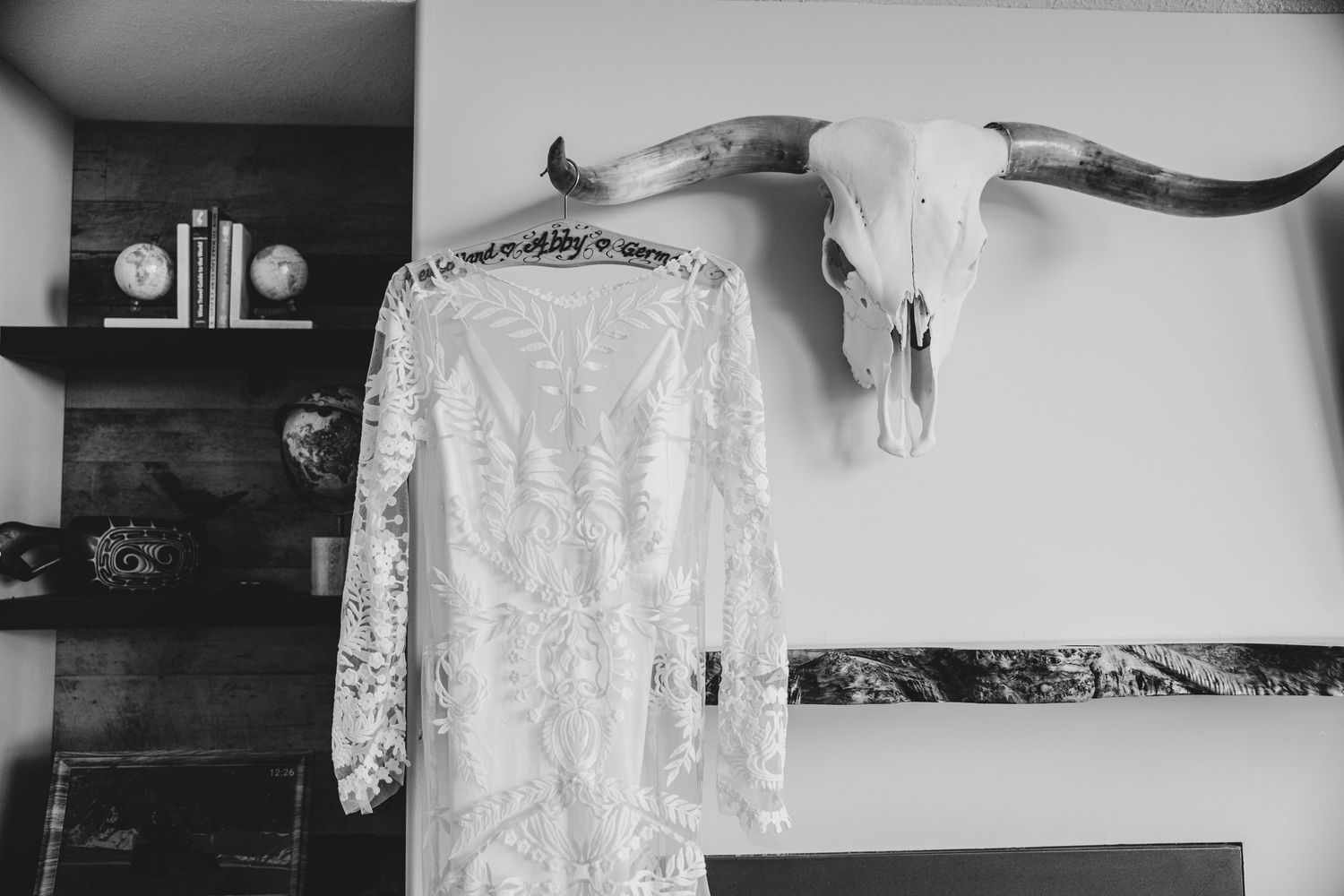 A white lace wedding dress hangs next to a mounted longhorn skull on a white wall.