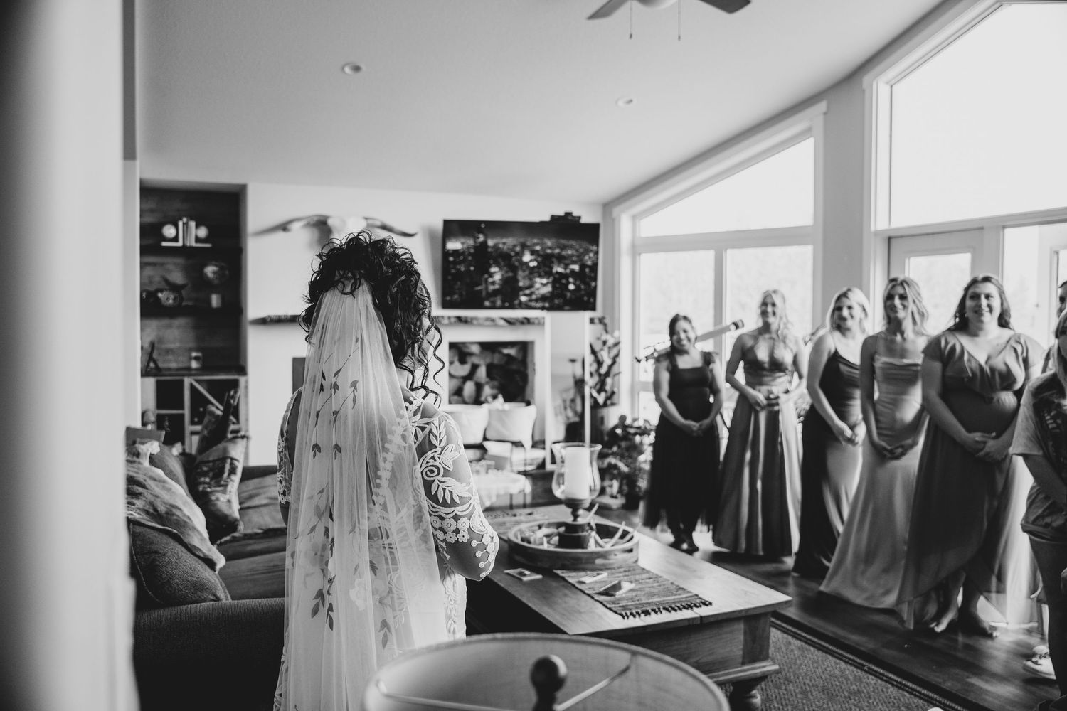 Black and white photo shows a wedding dress hanging in a bridal suite while bridesmaids gather in the background.