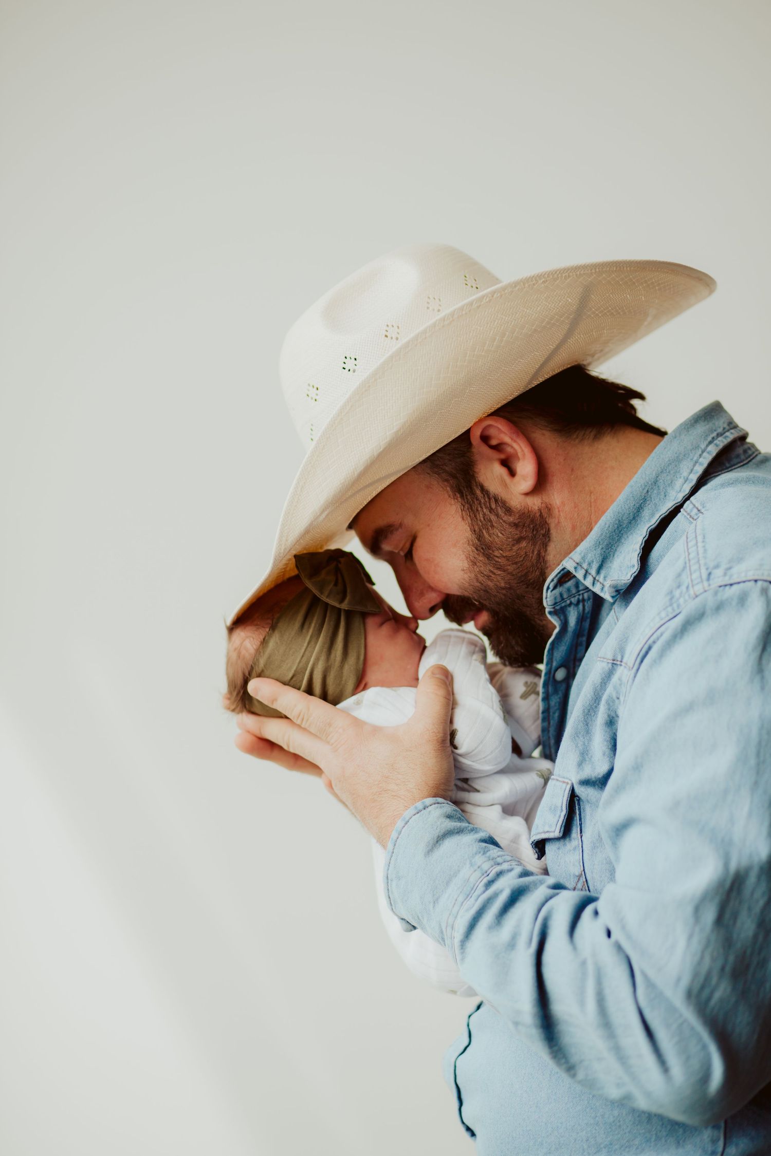 A person wearing a white cowboy hat and denim shirt holds and embraces a small baby close to their chest.