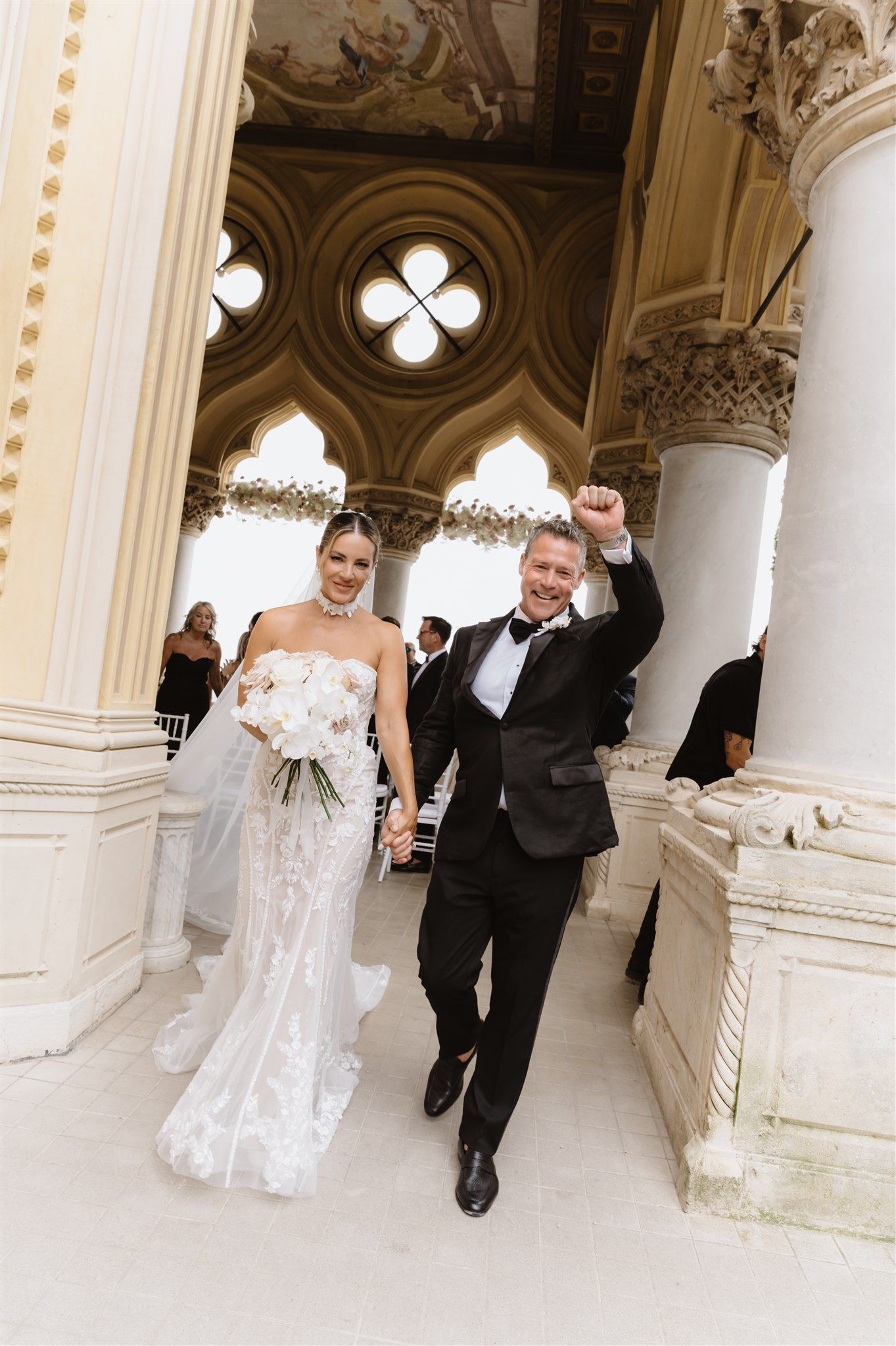 Bride and groom walking through an ornate hallway with gothic architecture and columns.