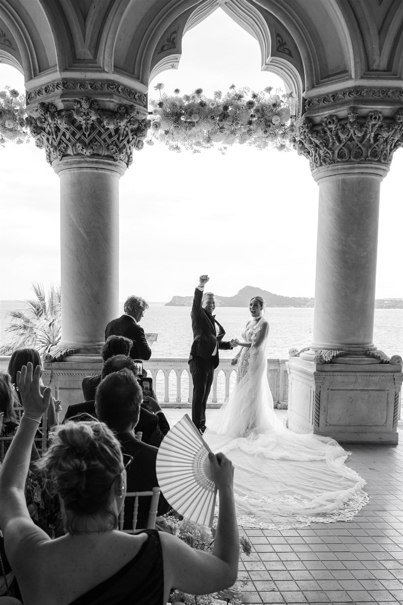 Wedding ceremony under ornate white columns with guests and ocean view.