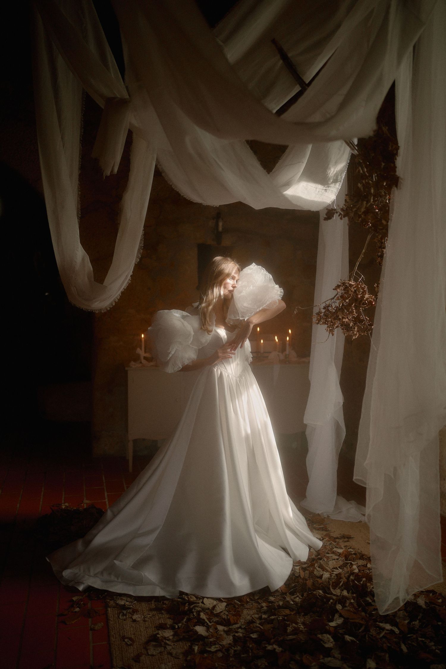 A ethereal figure in a flowing white dress stands surrounded by draped fabrics and candlelight in a romantic vintage setting.