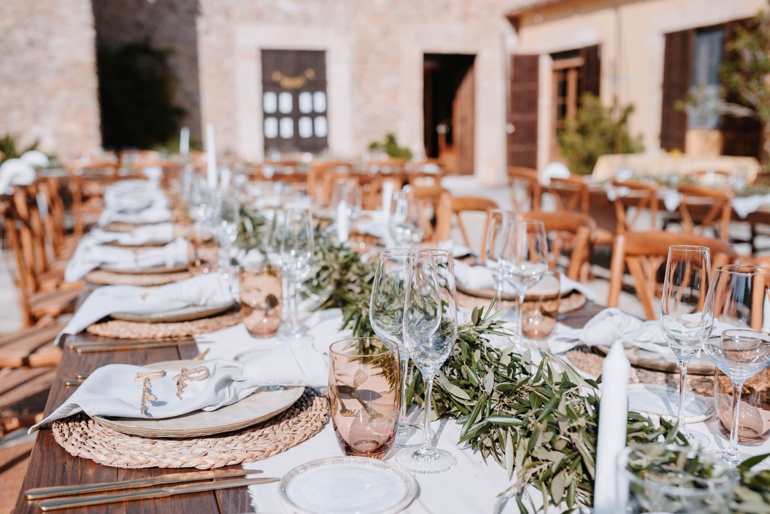 Elegante Hochzeitstische mit geflochtenen Platztellern, weißen Tellern und einer Girlande aus Olivenzweige als Tischdekoration auf Holz.