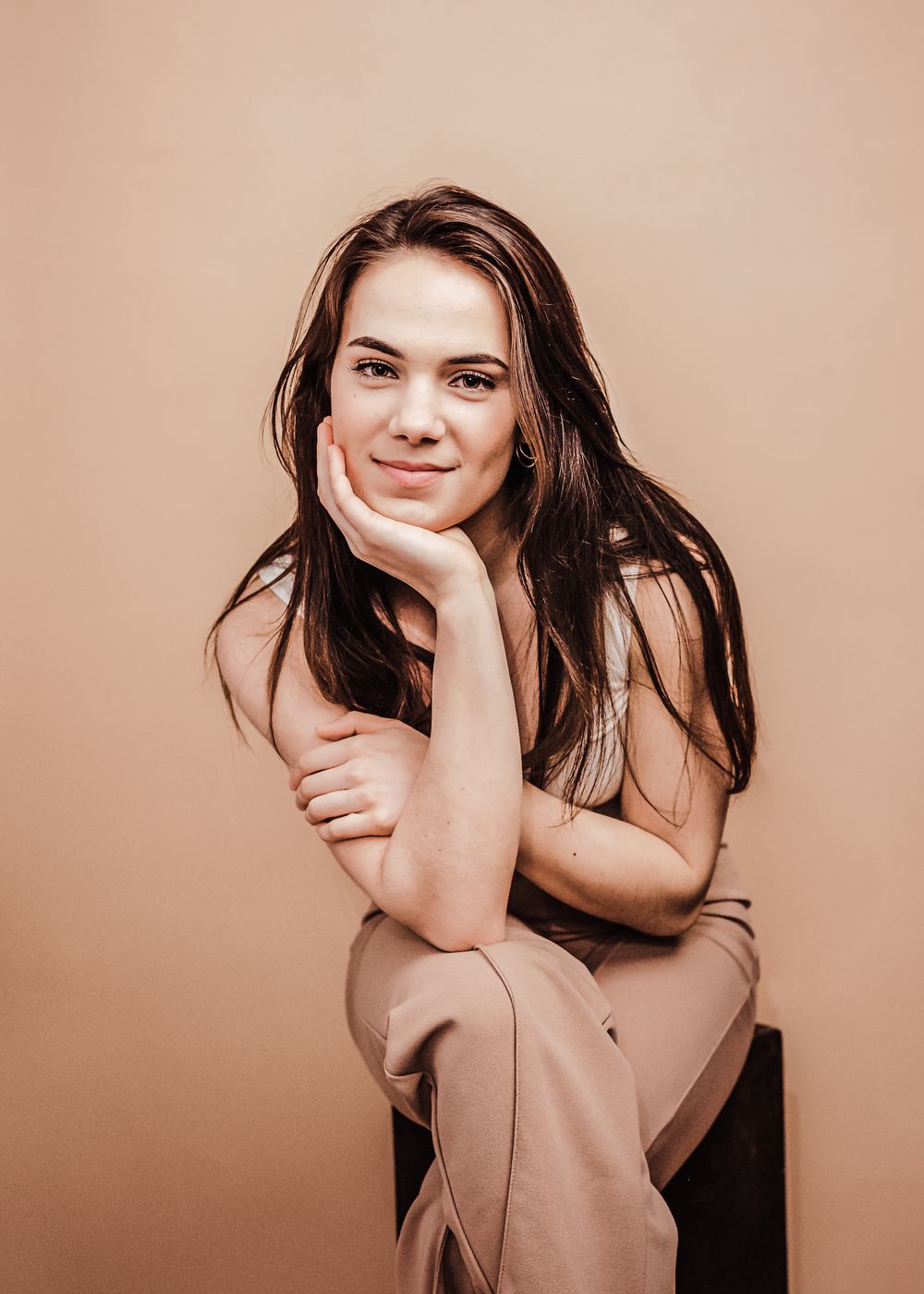 Portrait against beige background showing soft smile and long dark hair while resting chin on hand in casual pose.