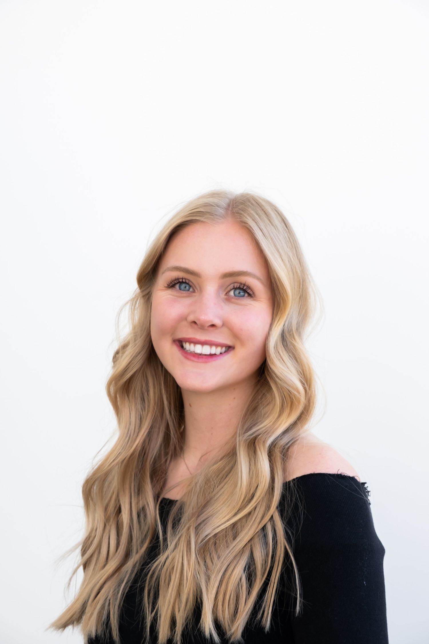 Person with wavy blonde hair smiling warmly against white background in black clothing.