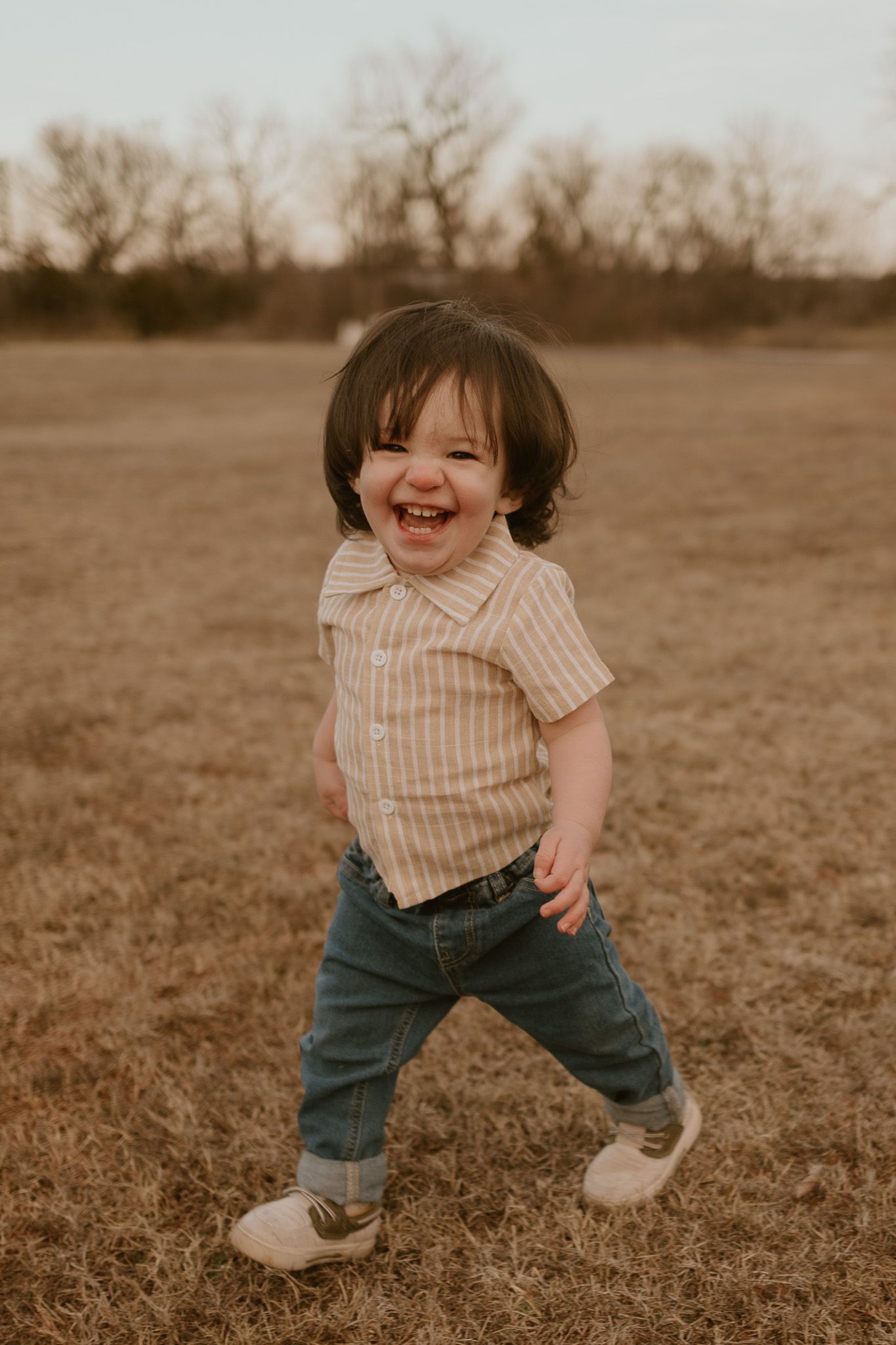 A small child walks through a brown field wearing a striped shirt and jeans while laughing joyfully.