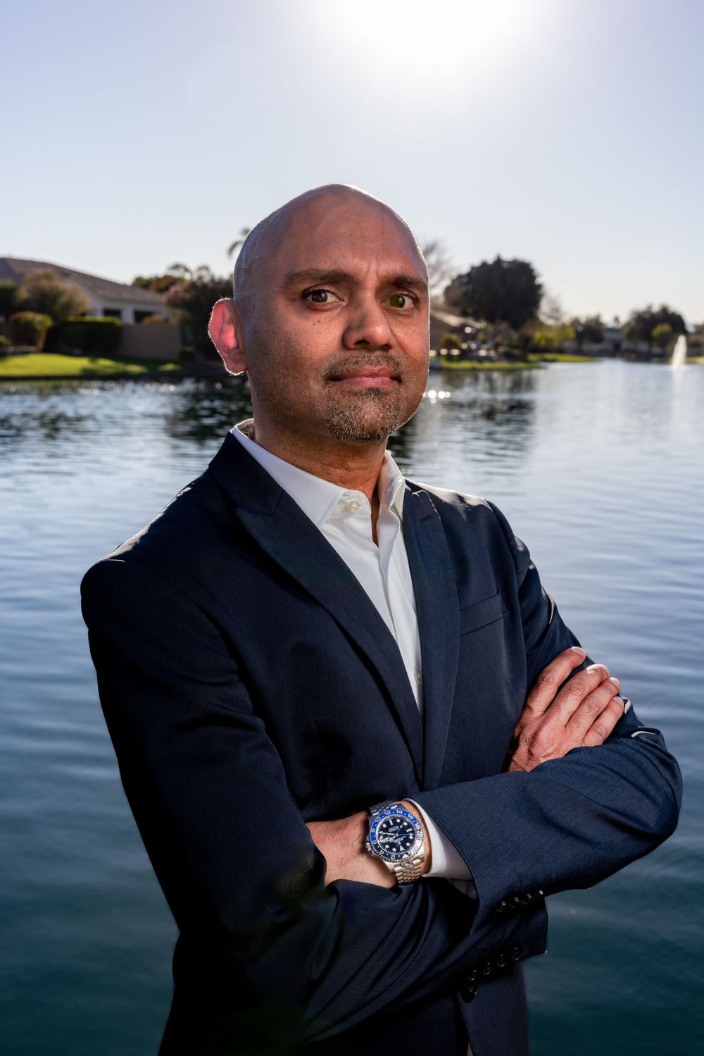 Man in navy blazer standing arms crossed near a serene lake with fountain in background.