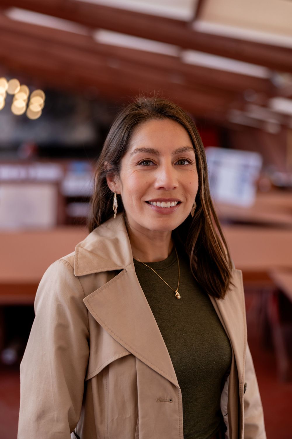 Smiling woman in beige trench coat and green top posing inside warm-lit restaurant.