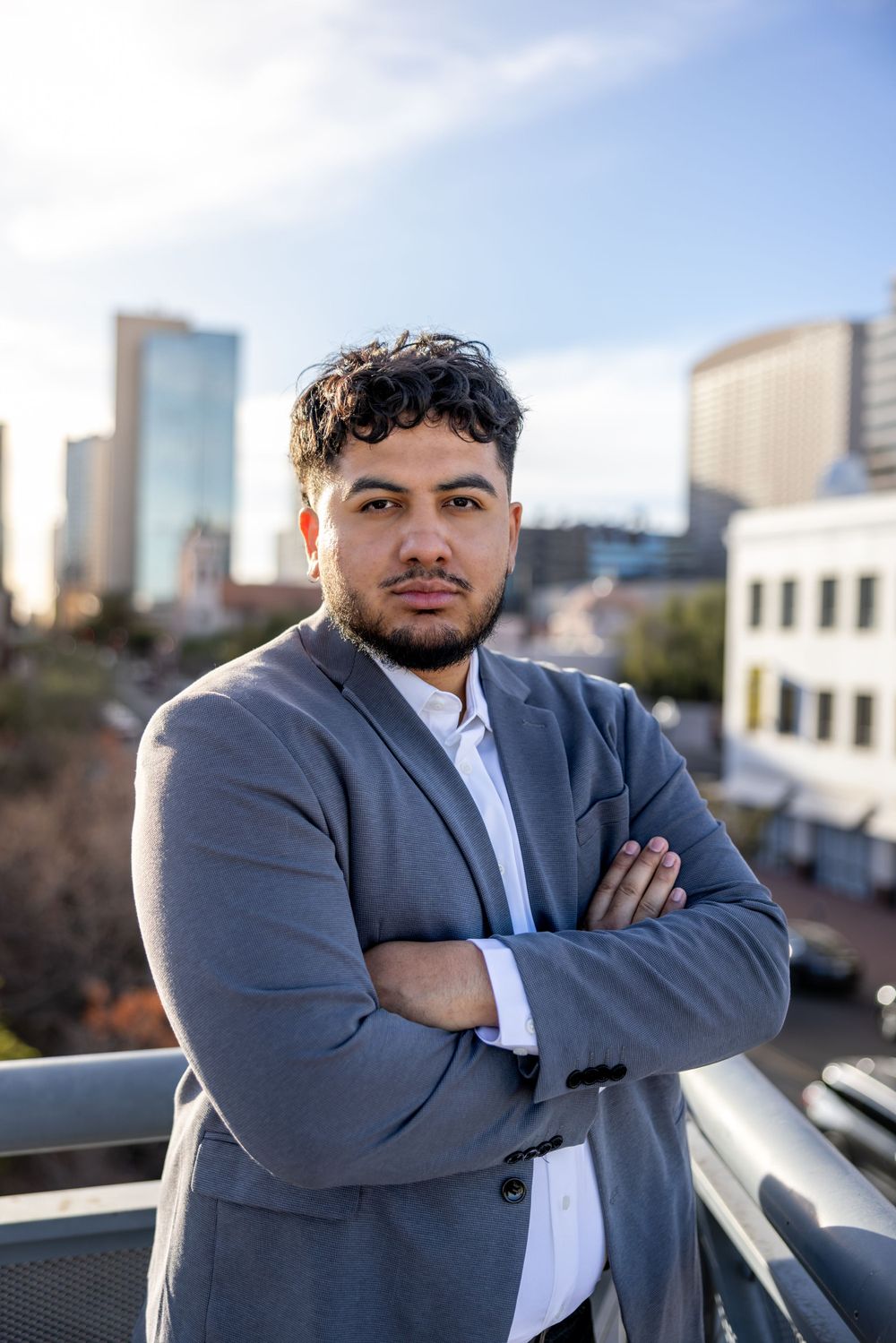 Young man in gray blazer standing with arms crossed on urban rooftop with city skyline behind him.