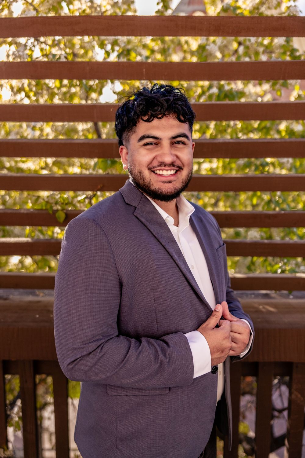 Smiling young man in gray suit jacket posing in front of wooden lattice fence with trees.