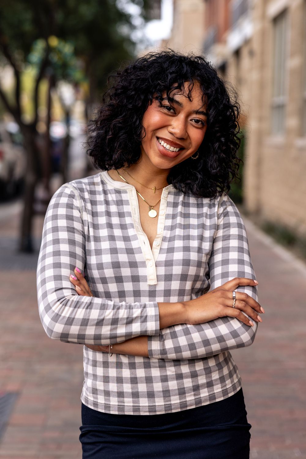 Smiling woman with curly hair in plaid top standing on a brick sidewalk with arms crossed.