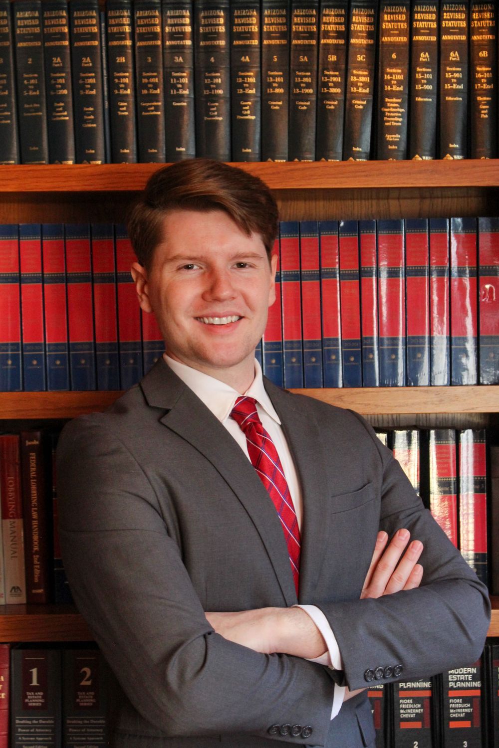 Smiling man in gray suit with arms crossed posing in front of a law library bookshelf.