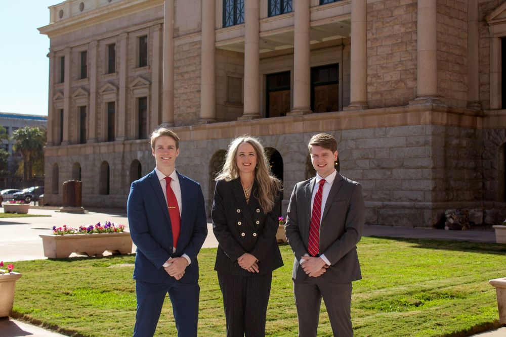 Three professionals in formal attire posing outside Arizona State Capitol building on sunny day.