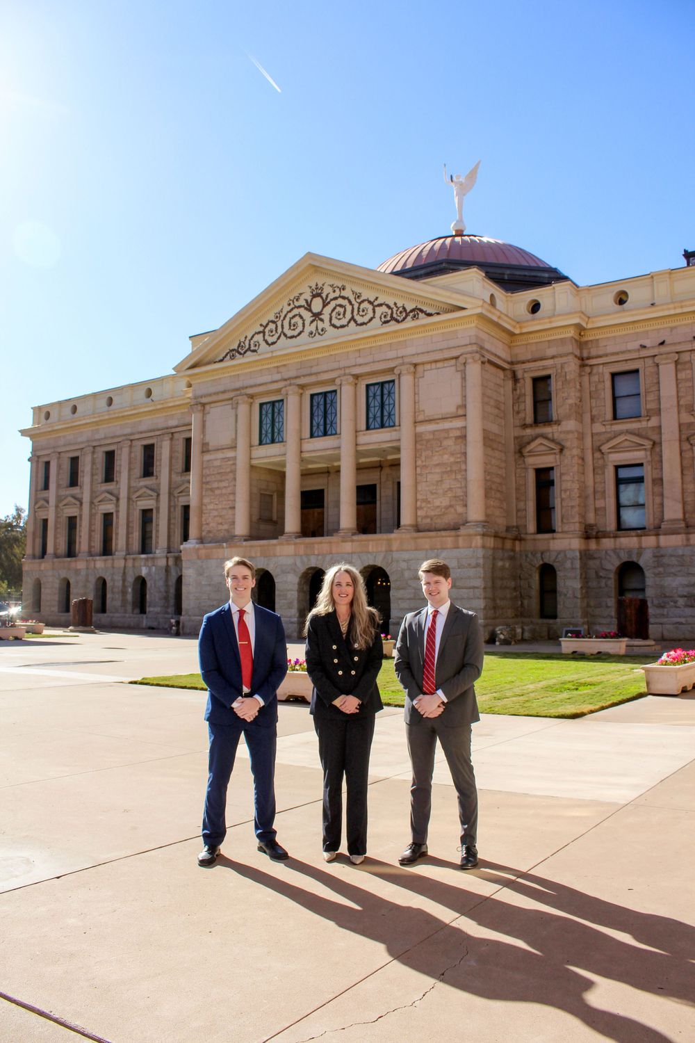 Three formally dressed young professionals stand together in front of the Arizona State Capitol building in bright sunlight.