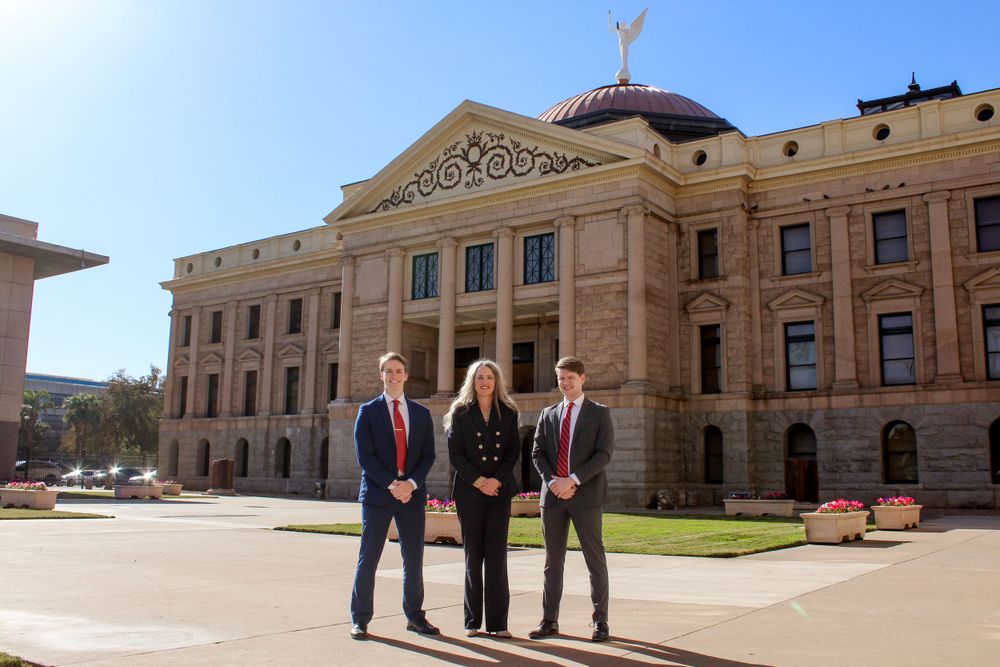 Three professionals in business attire pose in front of the ornate Arizona State Capitol building on a clear day.
