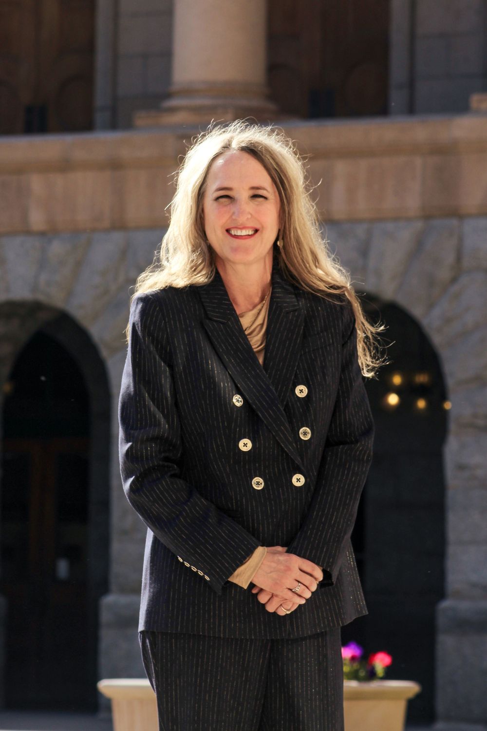 Professional woman in dark pinstripe suit smiling outdoors in front of a government building.