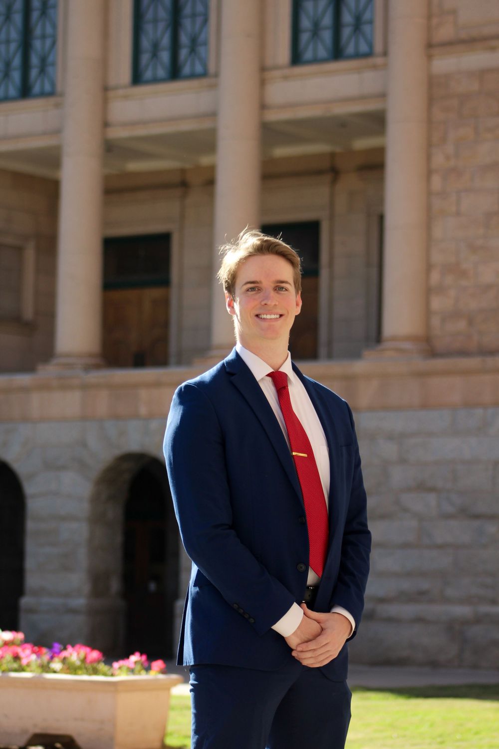 Smiling young man in navy blue suit and red tie posing outside a government building.