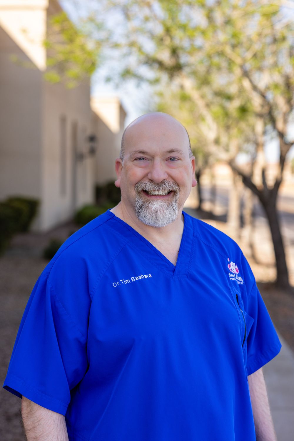 Smiling bald male doctor in blue scrubs standing outdoors near building entrance.