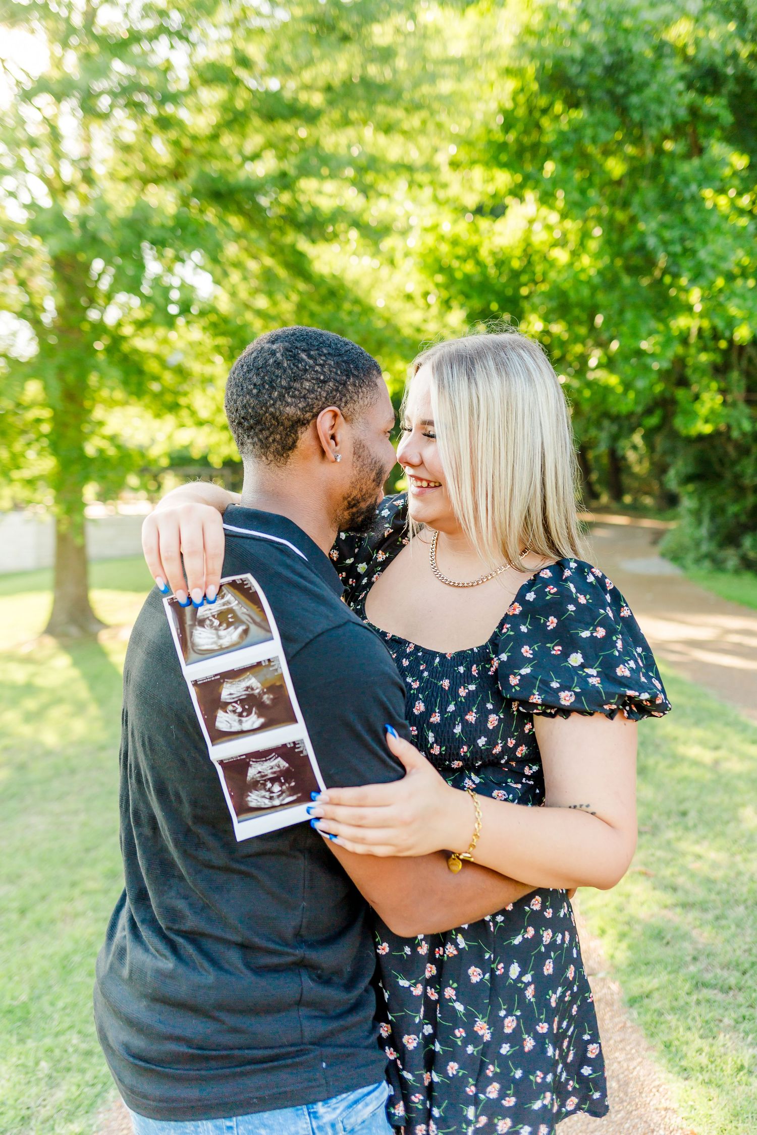 A couple embraces outdoors while holding ultrasound photos in a sunlit wooded setting during a pregnancy announcement.