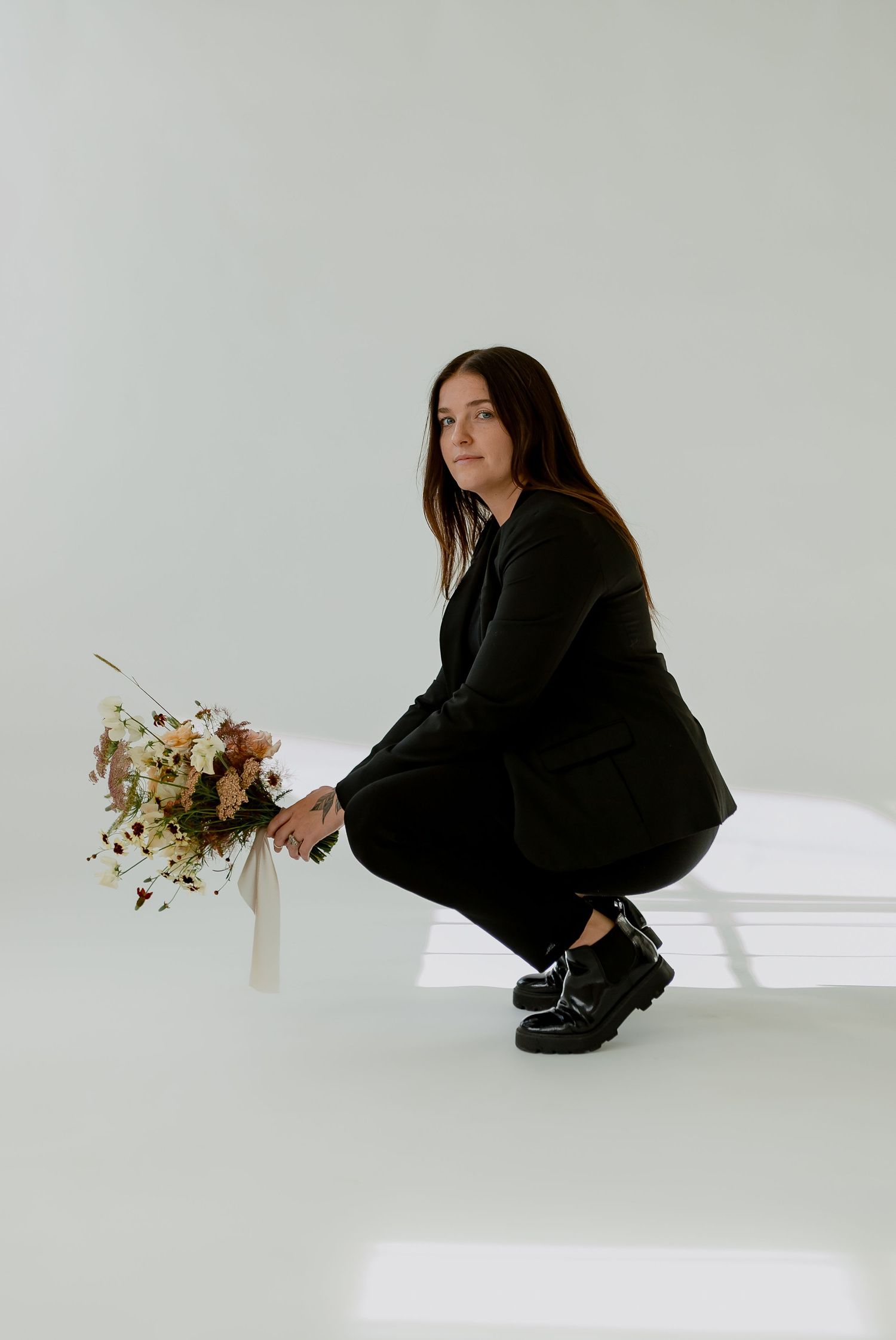 Person in black outfit crouching with floral arrangement against white studio backdrop.