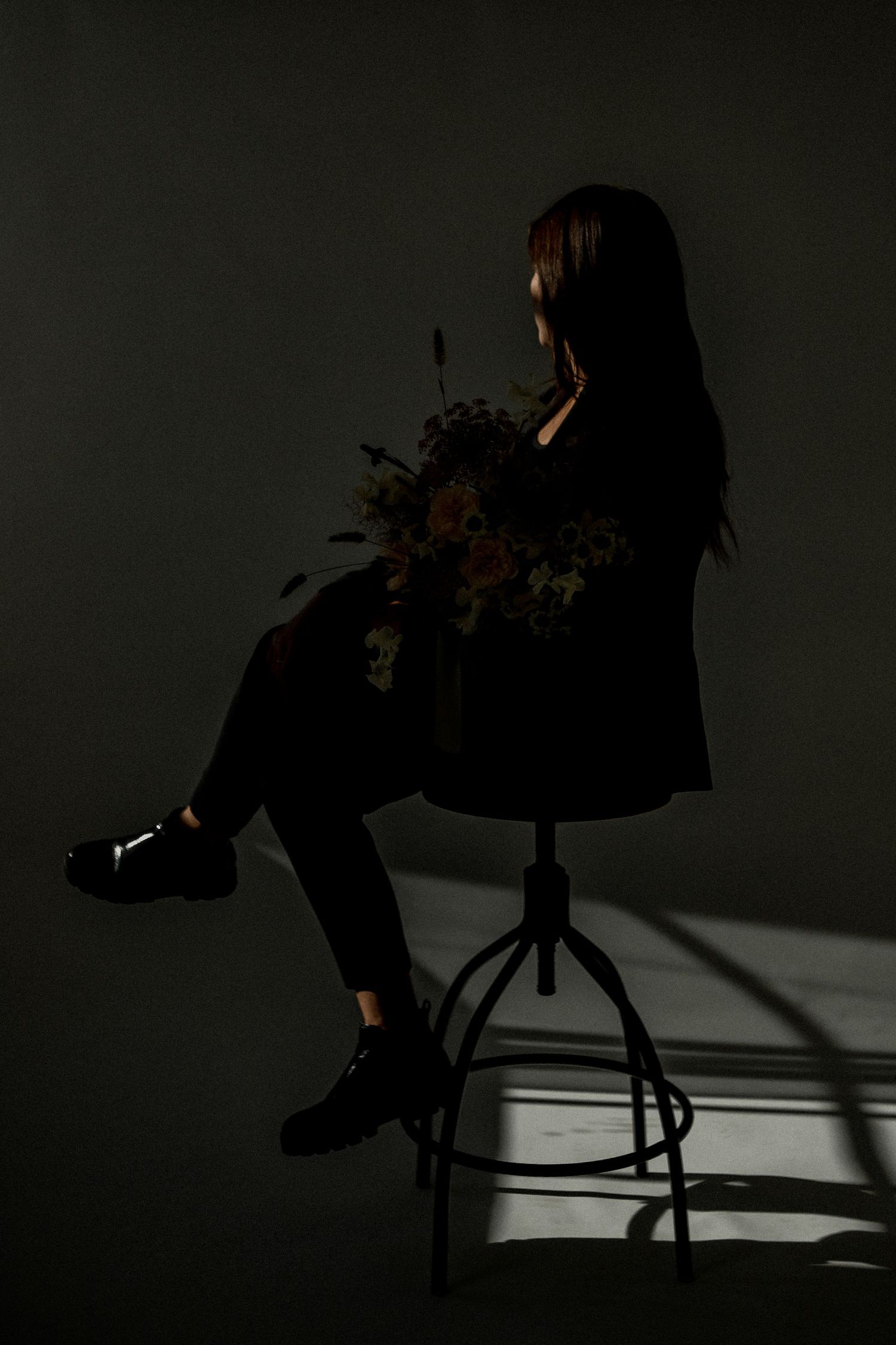 Moody artistic portrait of someone seated on a stool in dark clothing against a shadowy background.