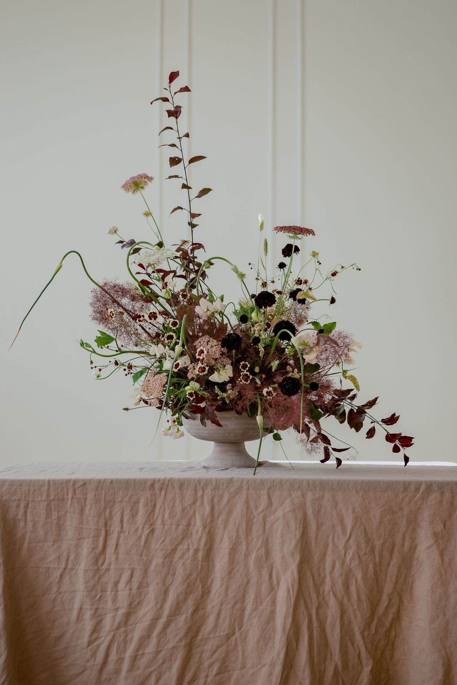 Elegant floral arrangement with pink and burgundy blooms in pottery vase on linen tablecloth.