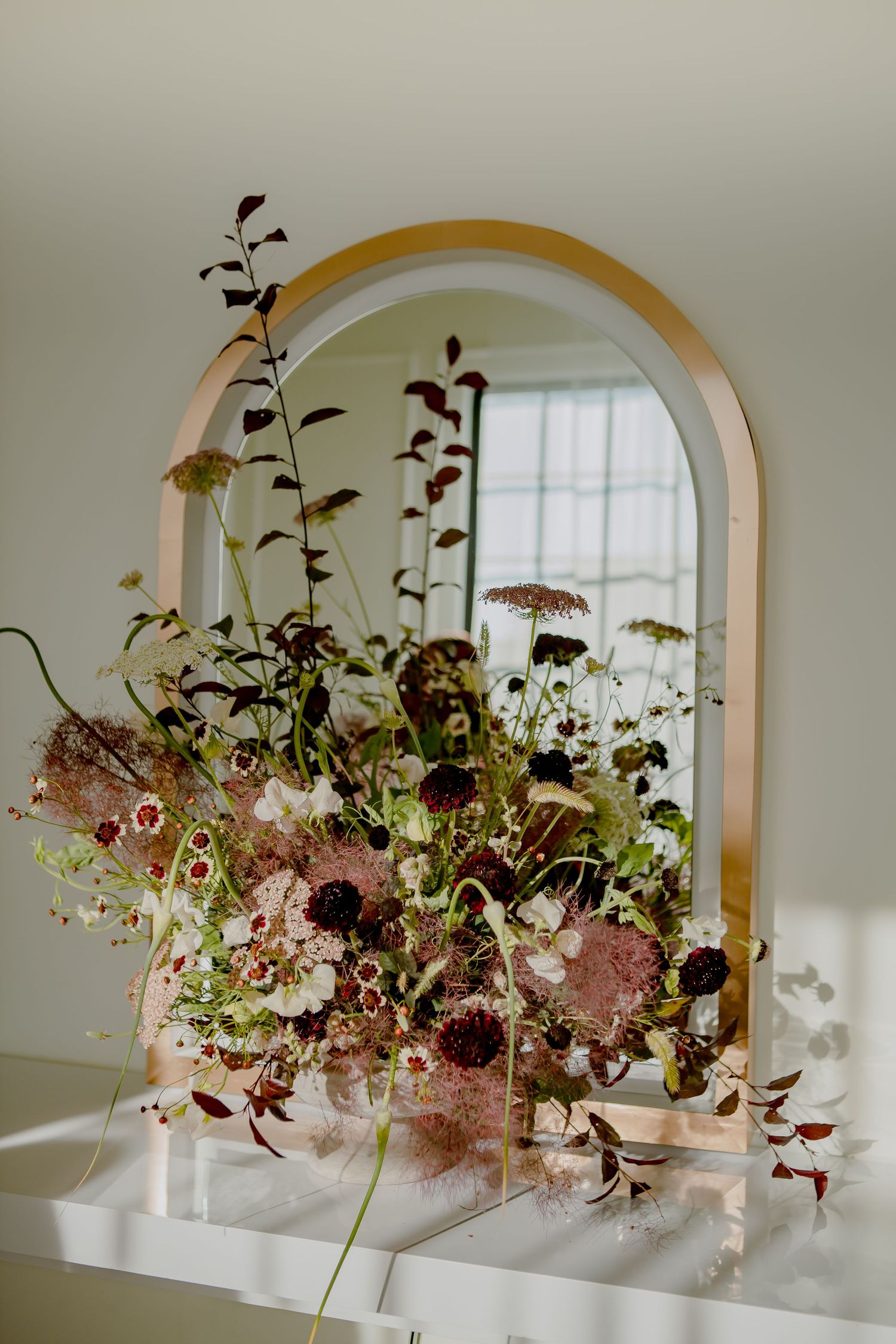 Floral arrangement reflected in arched golden mirror against white wall.