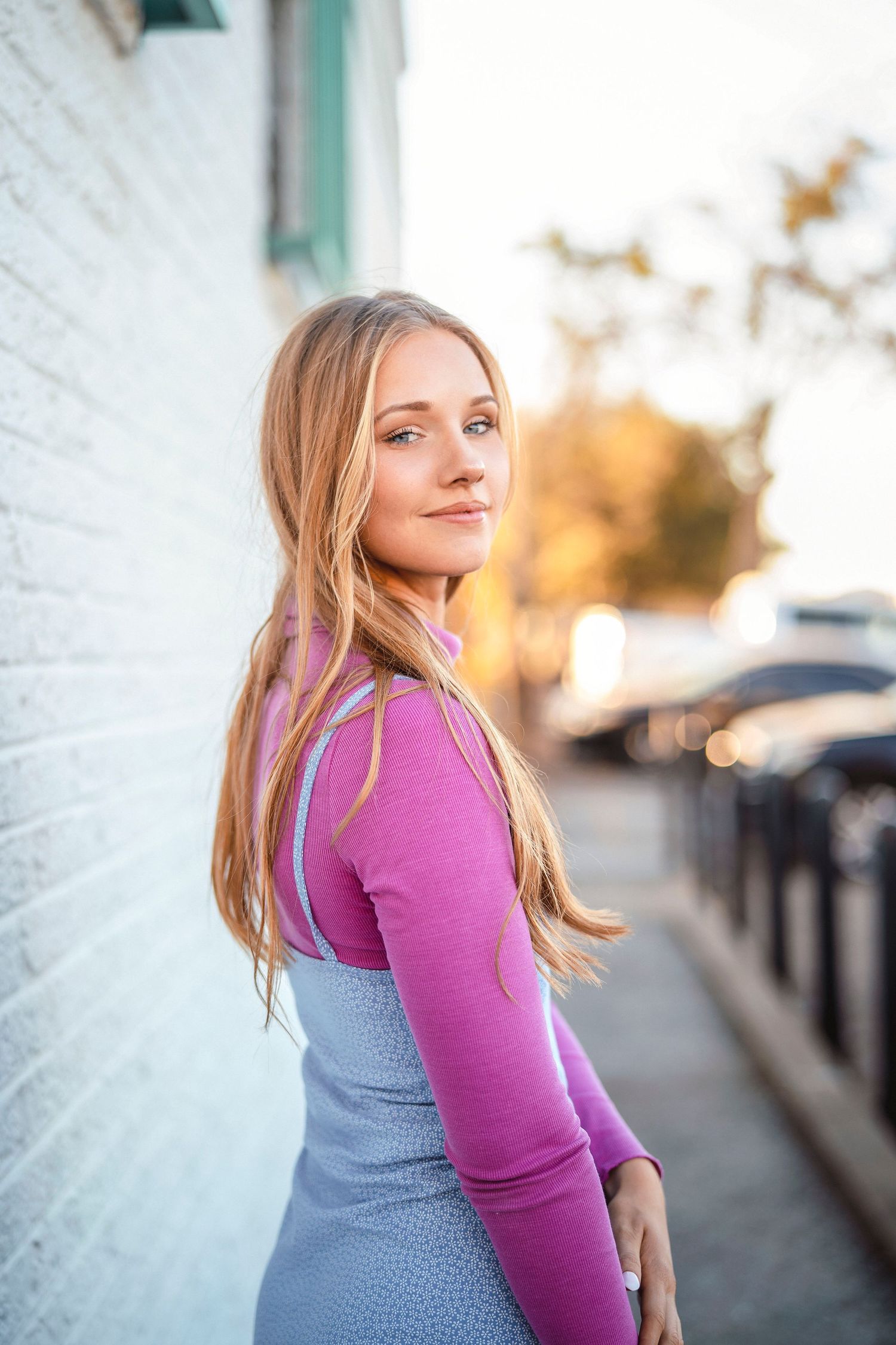 Woman in purple long sleeve top and blue dress poses against white wall in golden hour lighting.