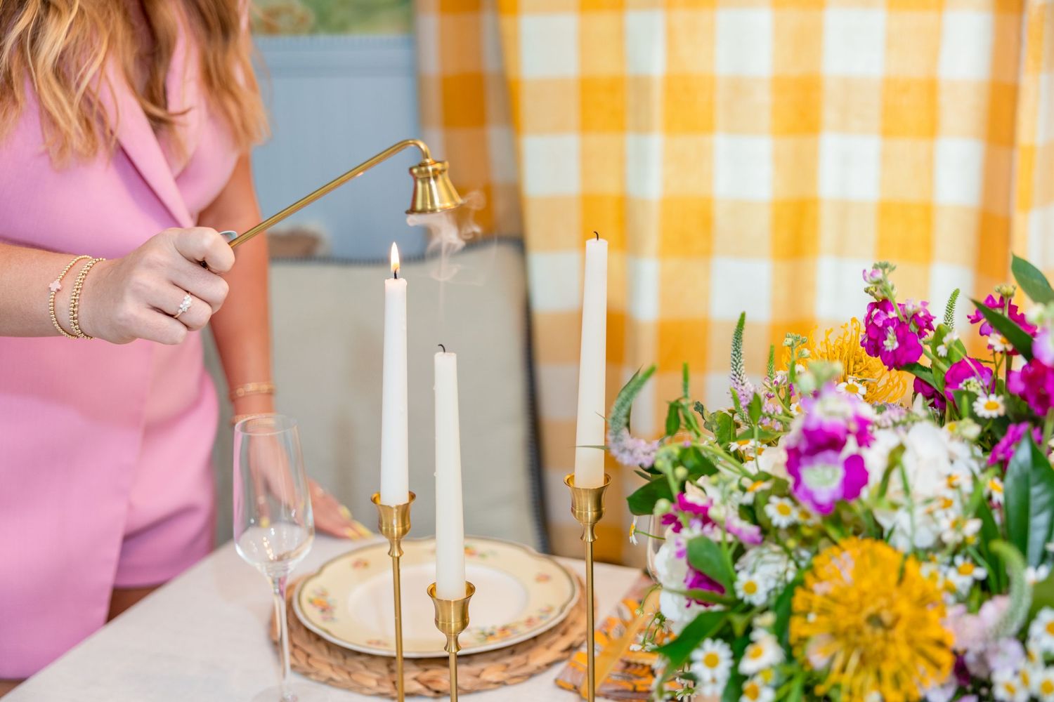 Woman in pink dress lighting white taper candles on a gold candleholder beside a colorful floral arrangement.