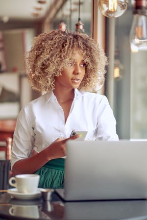 Woman with curly blonde hair works on laptop at cafe with coffee cup.