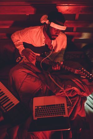 Musician working on music production with guitar and laptop under red lighting.
