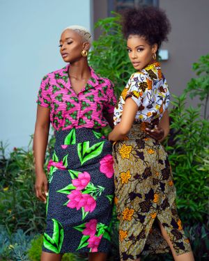Two models showcase vibrant African print dresses with bold floral patterns against a garden backdrop.