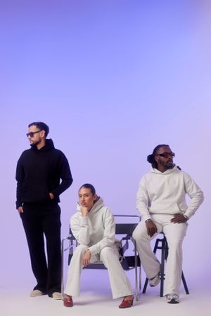 Three people in casual wear posing on chairs against purple gradient background.