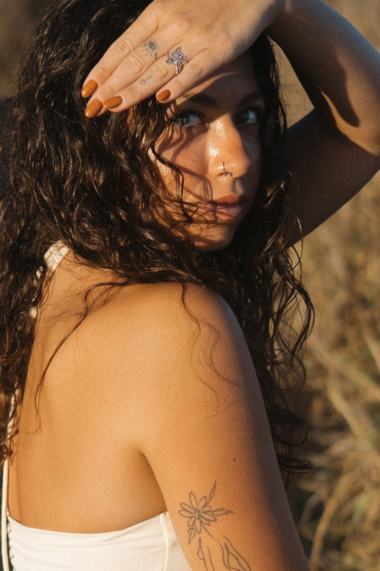 Portrait with long wet dark hair against golden sunlight, wearing white top with shoulder tattoo and ring.
