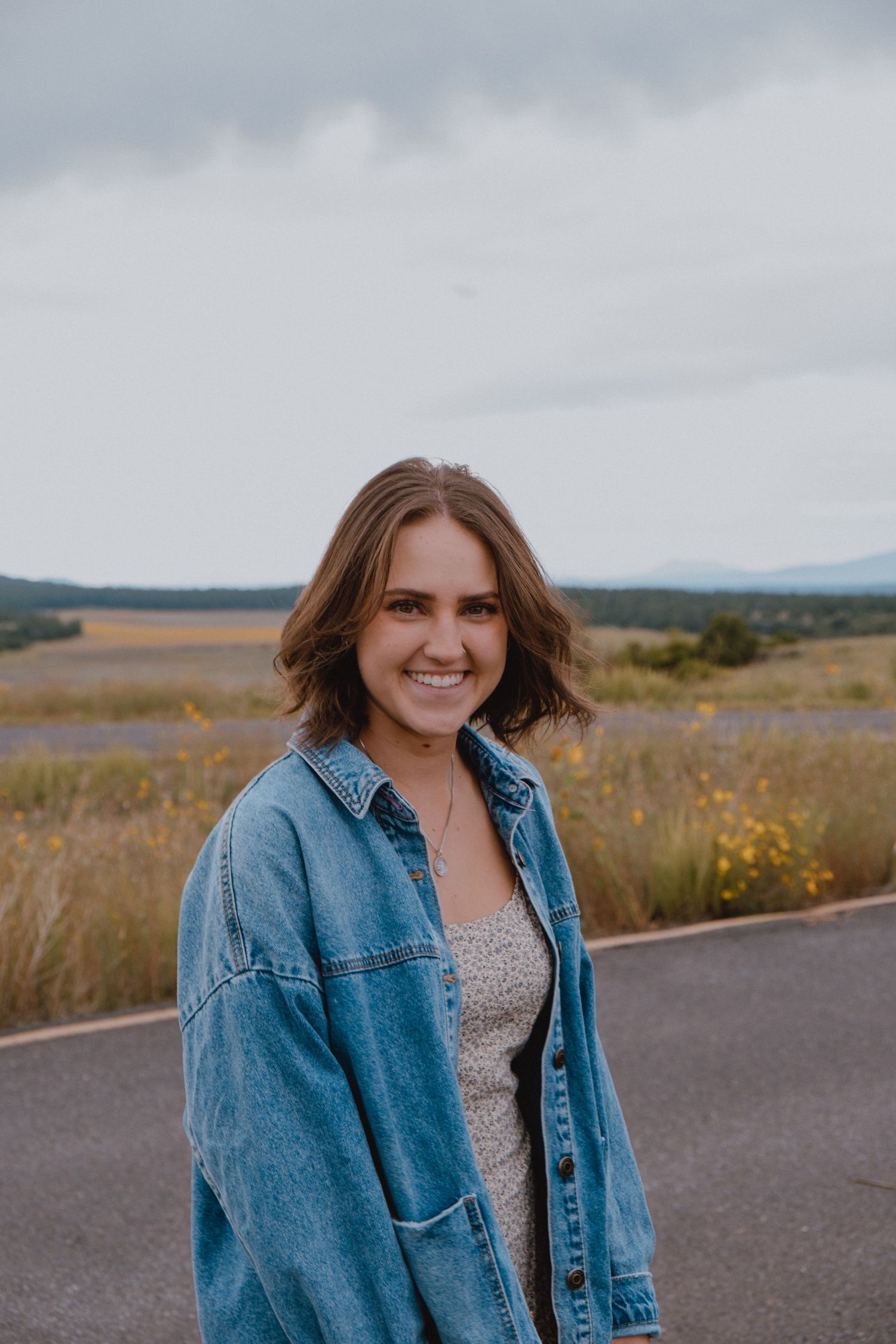 A person wearing a denim jacket stands on a road with mountains and meadows in the cloudy background.