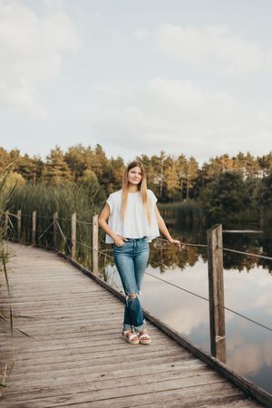 Young woman in white top and jeans smiling on a wooden dock by a calm lake surrounded by lush green trees.