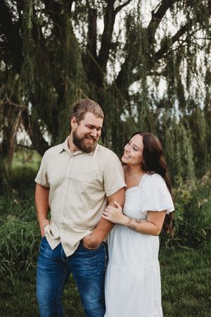 A bearded man in a beige shirt and a woman in a white dress smile together outdoors near a willow tree.