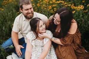 Candid moment of parents and young child laughing together in a field of yellow wildflowers at sunset.