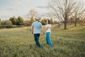 A couple holds hands walking through a lush green field at golden hour near a bare tree.
