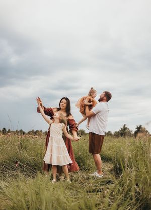 Family of four playing in a grassy field, parents lifting children under cloudy skies.