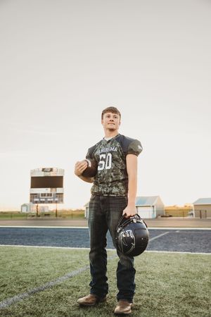 A young football player stands on a field wearing a jersey number 50 and holding a helmet at sunset.