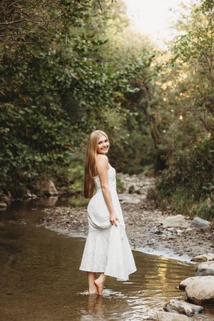 A person in a white dress stands playfully in a shallow creek surrounded by trees during golden hour.