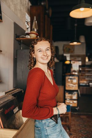 Person in red sweater and jeans smiles while sitting in a cozy coffee shop interior with warm lighting.