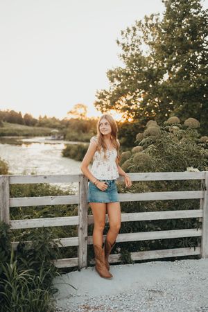 Young woman in floral top, denim skirt, and cowboy boots leaning on a wooden fence at golden hour.