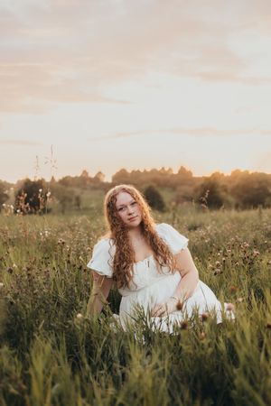 Young woman with curly red hair in white dress sitting in wildflower meadow during golden sunset.